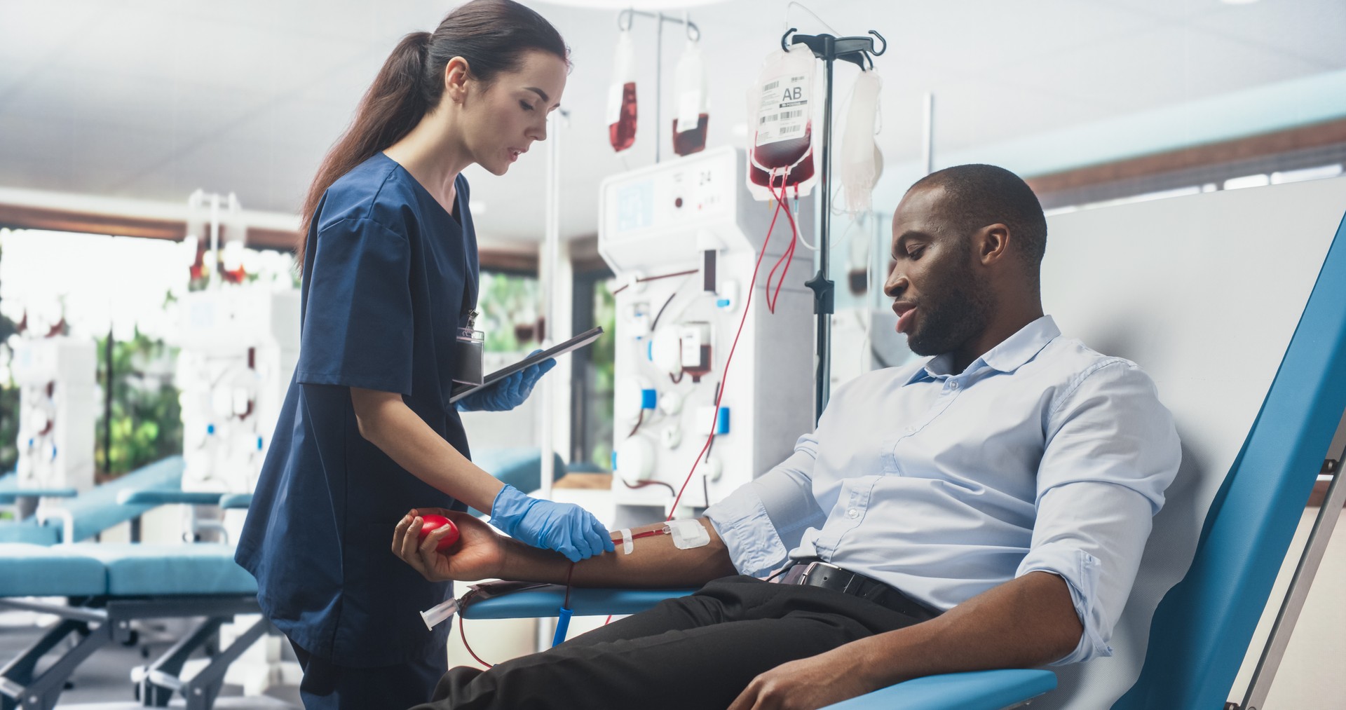 Black Businessman Donating Blood For People In Need In Bright Hospital. Female Nurse With Tablet Computer Coming In To Check Progress And Well-Being Of Donor. Donation For Heart Surgery Patients.